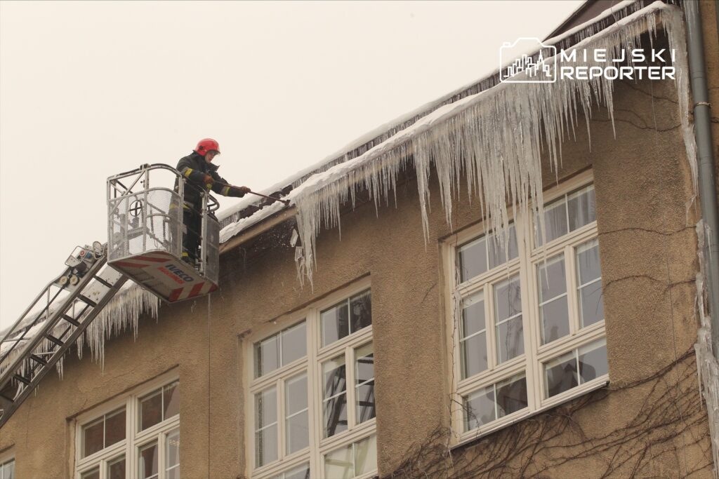 Strażak w kasku i mundurze usuwa sople lodu z dachu budynku przy użyciu podnośnika.