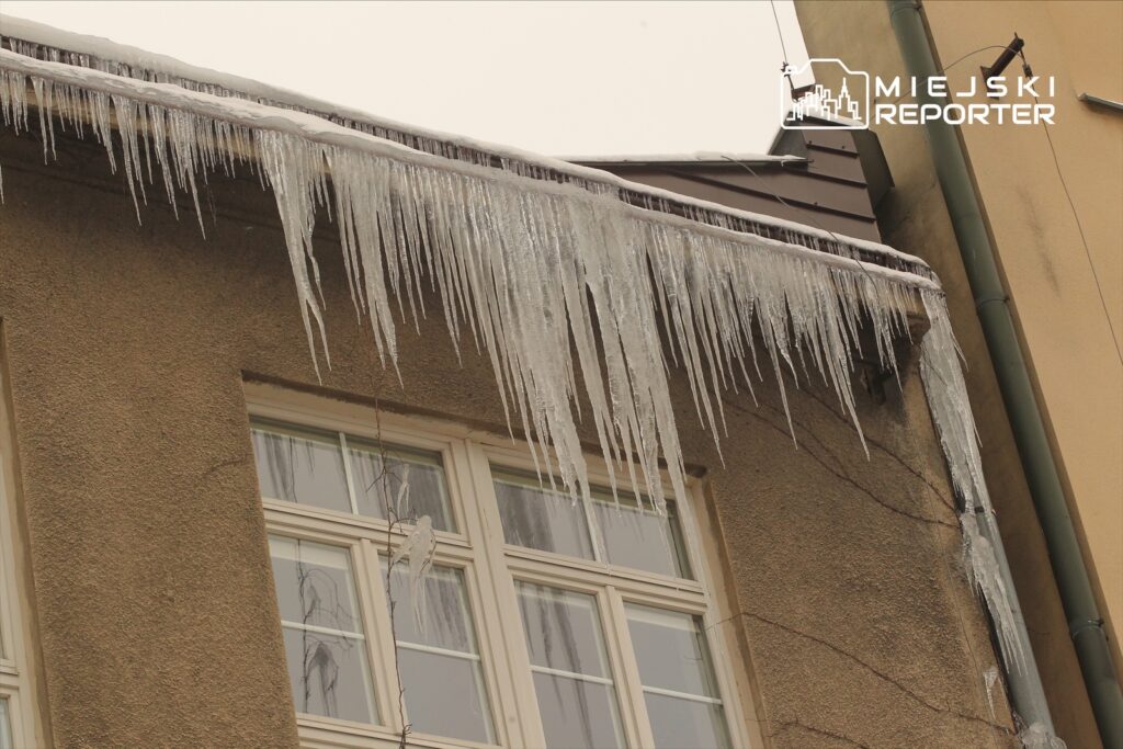 Sople lodu zwisające z rynny budynku, w tle okna z białymi ramami i szaro-beżowa elewacja.
