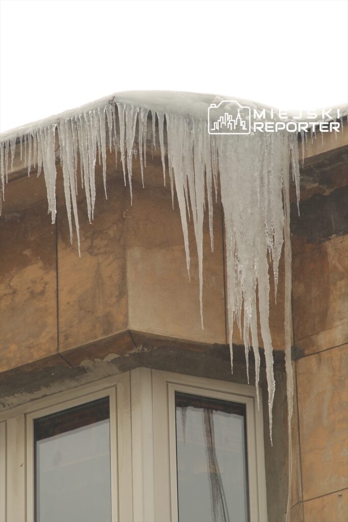 Sople lodu zwisające z krawędzi dachu nowoczesnego budynku o szarej elewacji.