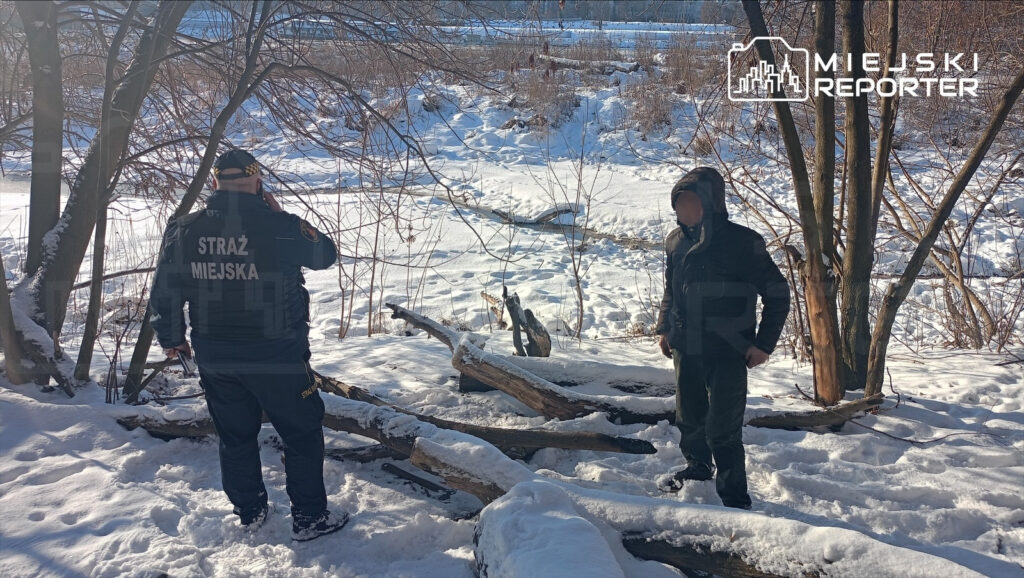 Mężczyzna w mundurze straży miejskiej rozmawia przez telefon, stojąc obok drugiego mężczyzny na zaśnieżonym brzegu rzeki.