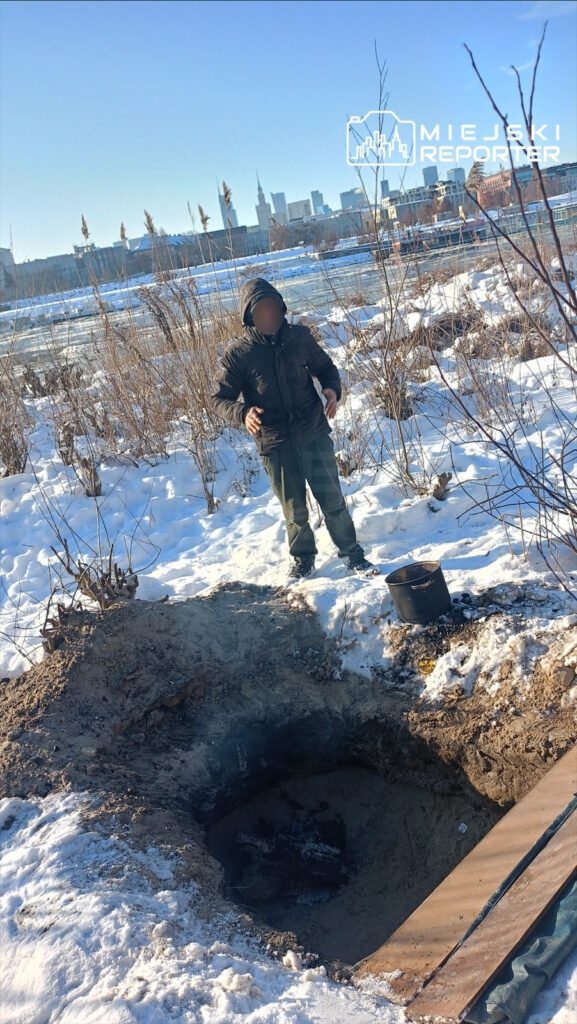 Chłopiec w ciemnej kurtce stoi obok wykopanej dziury w śniegu, obok niego czarny garnek z resztkami ognia. W tle widać miasto.