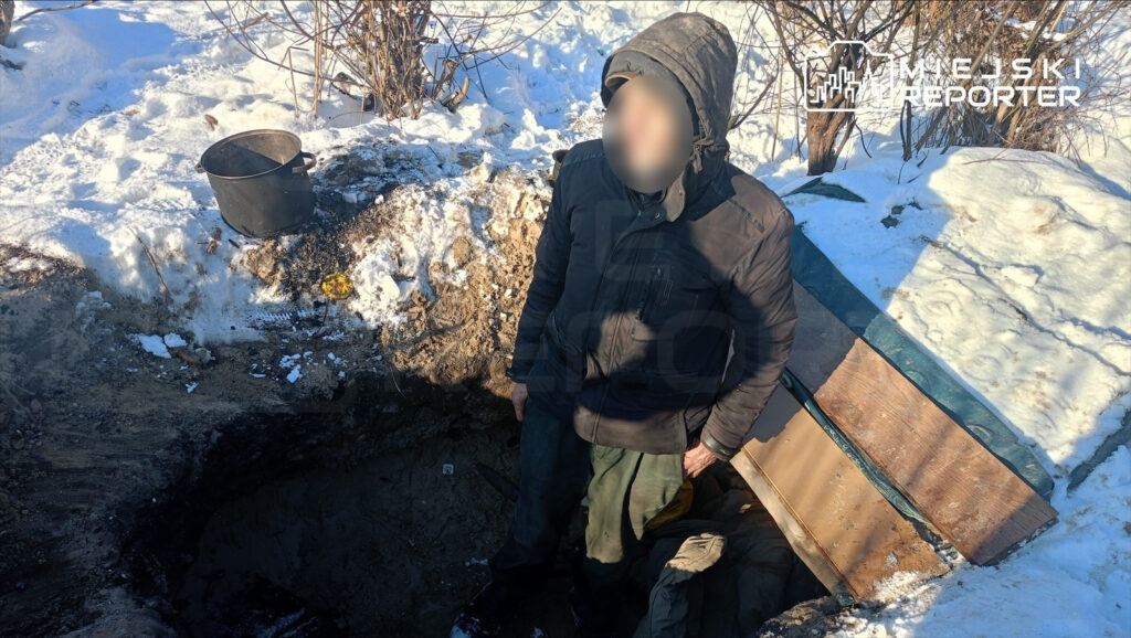 Mężczyzna w ciemnej kurtce stoi obok wykopu w zaśnieżonym terenie, obok niego czarny garnek.