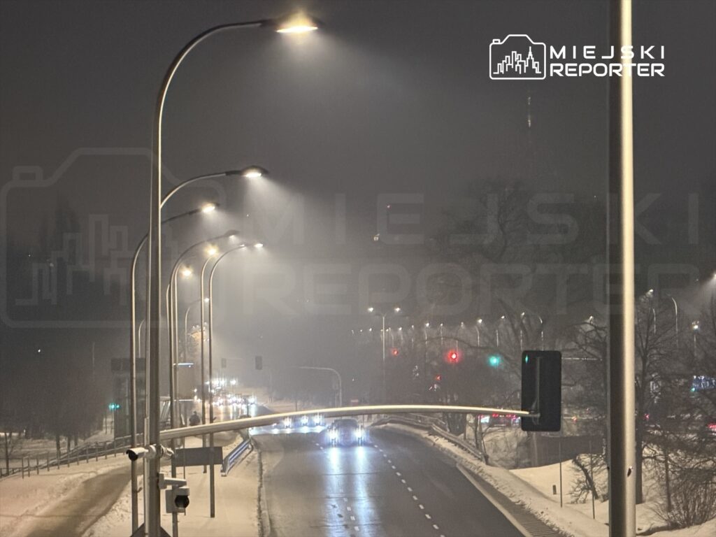 Oświetlona ulica w nocy z widocznymi latarniami, pojazdami i sygnalizacją świetlną w tle.