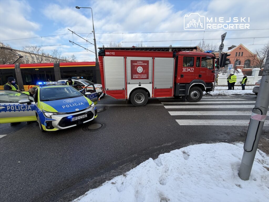 Na skrzyżowaniu zatrzymane pojazdy policyjne i wóz strażacki, w tle widoczne osoby w kamizelkach odblaskowych.