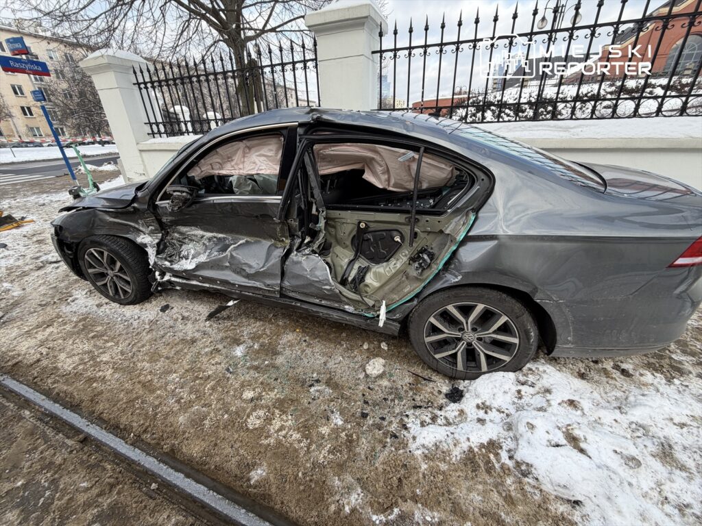 Uszkodzony samochód osobowy z wgniecioną lewą stroną zaparkowany na zaśnieżonej ulicy obok ogrodzenia.