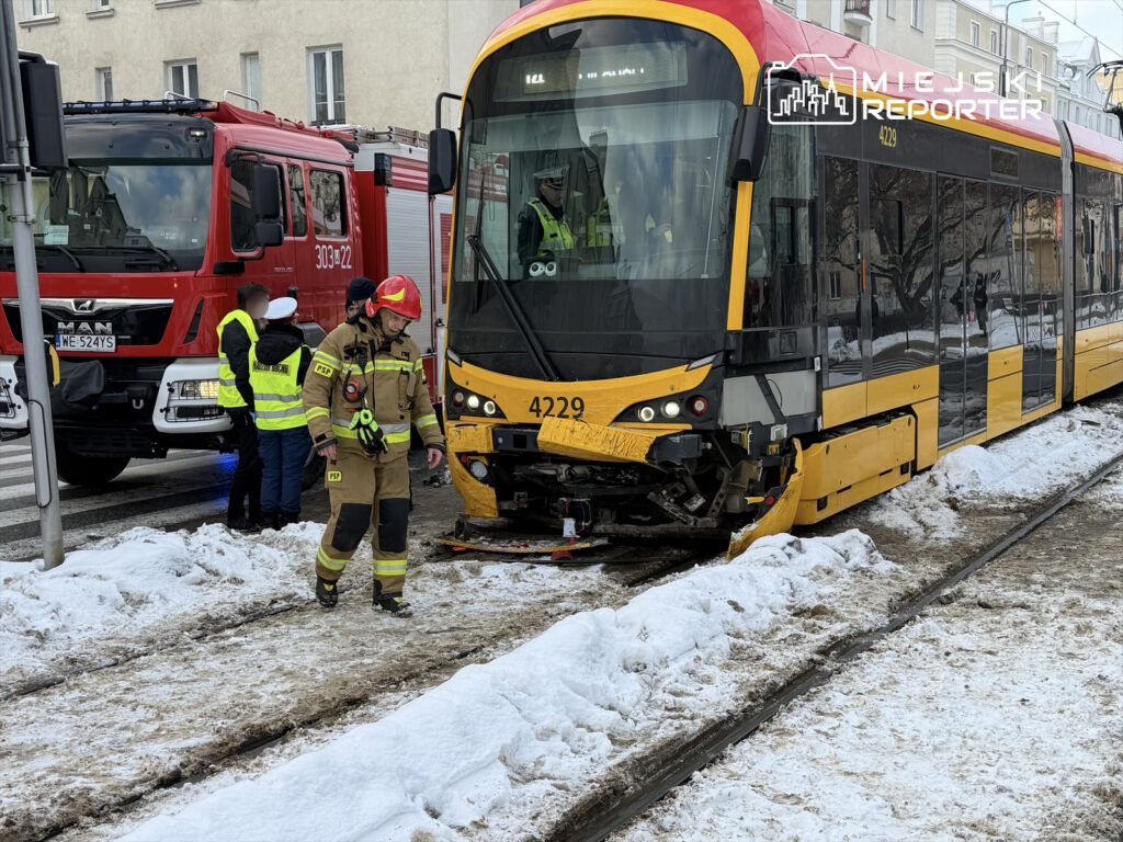 Strażak w mundurze bada uszkodzony tramwaj na śnieżnej ulicy, obok stoi wóz strażacki.