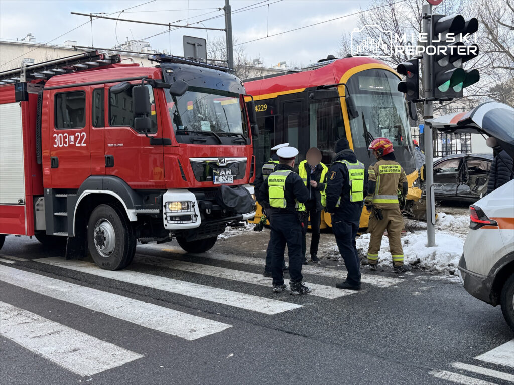 Czerwony wóz strażacki stoi obok uszkodzonego tramwaju, a na miejscu zdarzenia pracują policjanci i strażacy.