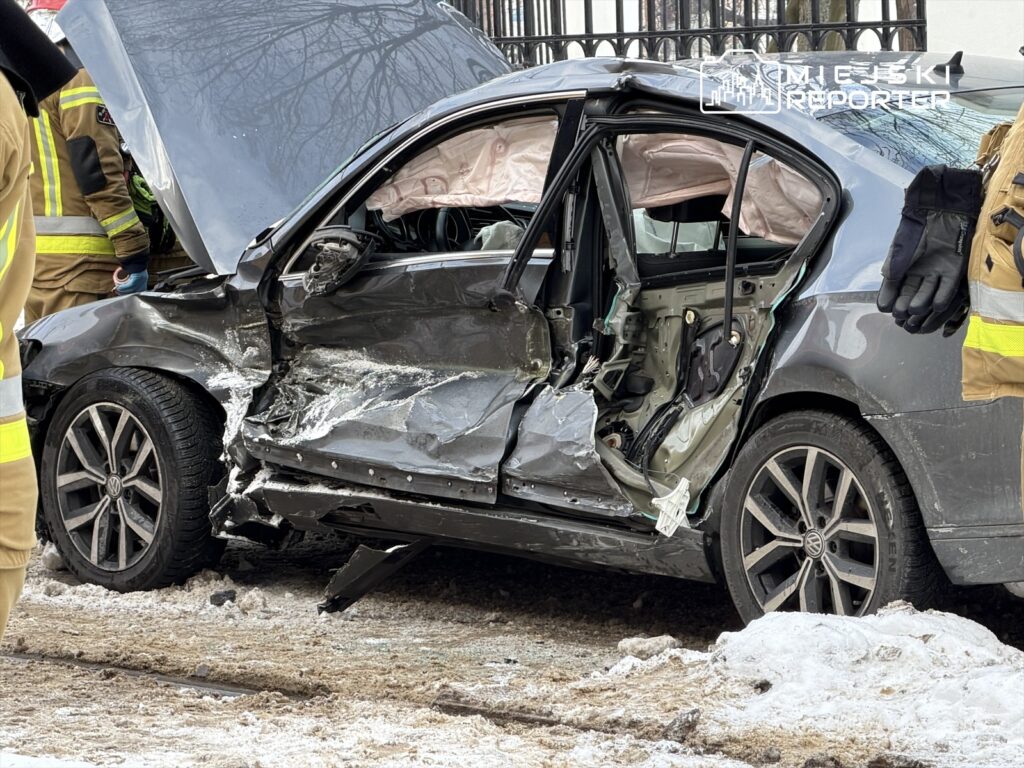 Uszkodzony samochód osobowy z otwartą maską, otwarta strona drzwi z widocznymi uszkodzeniami, w tle strażacy w mundurach.