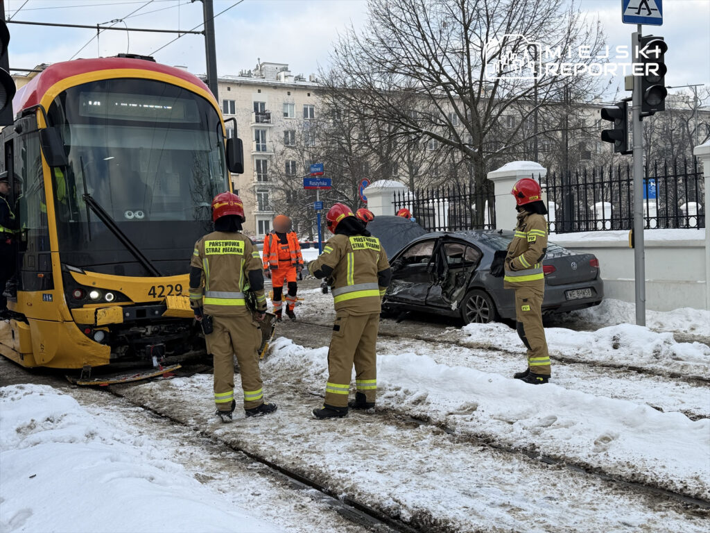 Ratownicy w mundurach, z hełmami, udzielają pomocy przy wypadku tramwajowym obok uszkodzonego samochodu na zaśnieżonej ulicy.
