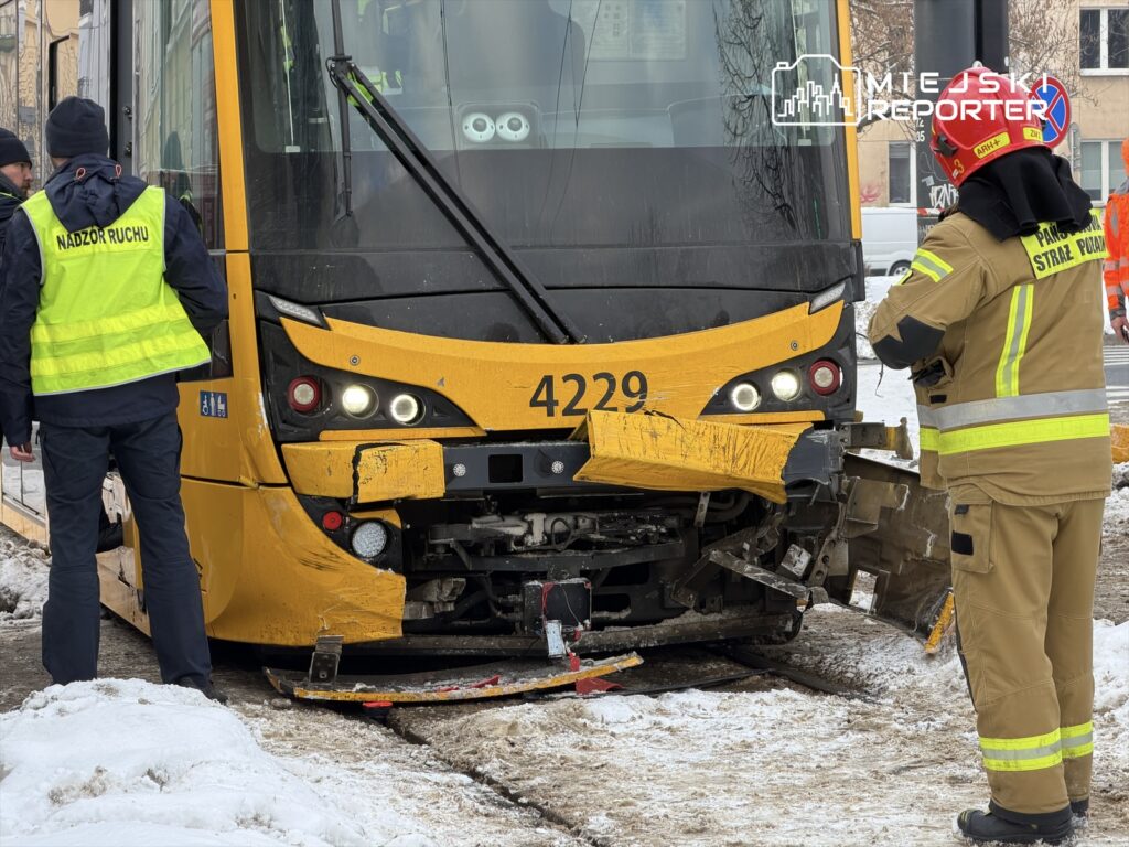 Grupa pracowników w odblaskowych kamizelkach oraz strażak w mundurze bada uszkodzony tramwaj na zaśnieżonej ulicy.