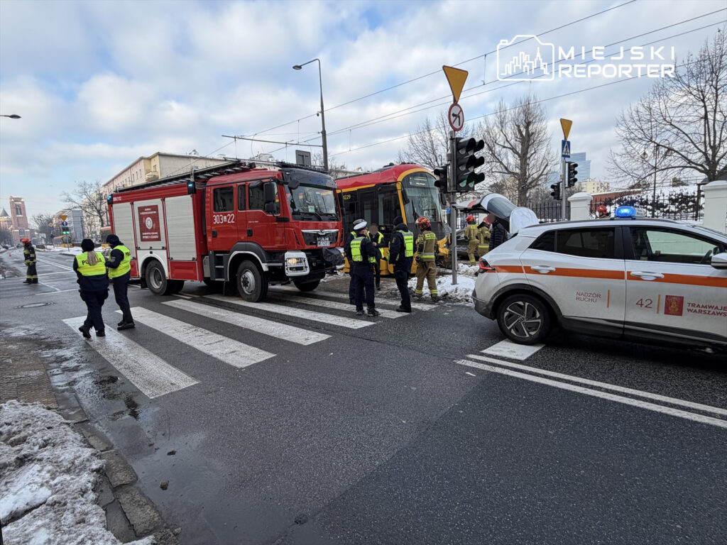 Grupa strażaków i policjantów pracuje przy wypadku drogowym, obok czerwonego wozu strażackiego i białego samochodu z sygnalizacją.