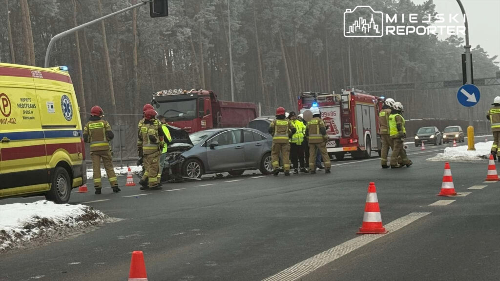 Grupa strażaków w mundurach i hełmach omawia sytuację przy wypadku drogowym, obok uszkodzonego samochodu.