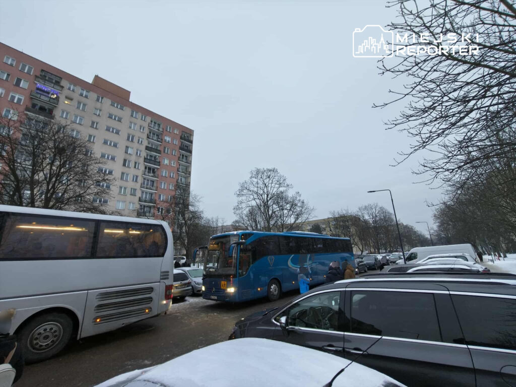 Autobus w niebieskim kolorze zatrzymany na zaśnieżonej ulicy, obok białego autokaru i zaparkowanych samochodów. W tle widać blok mieszkalny.