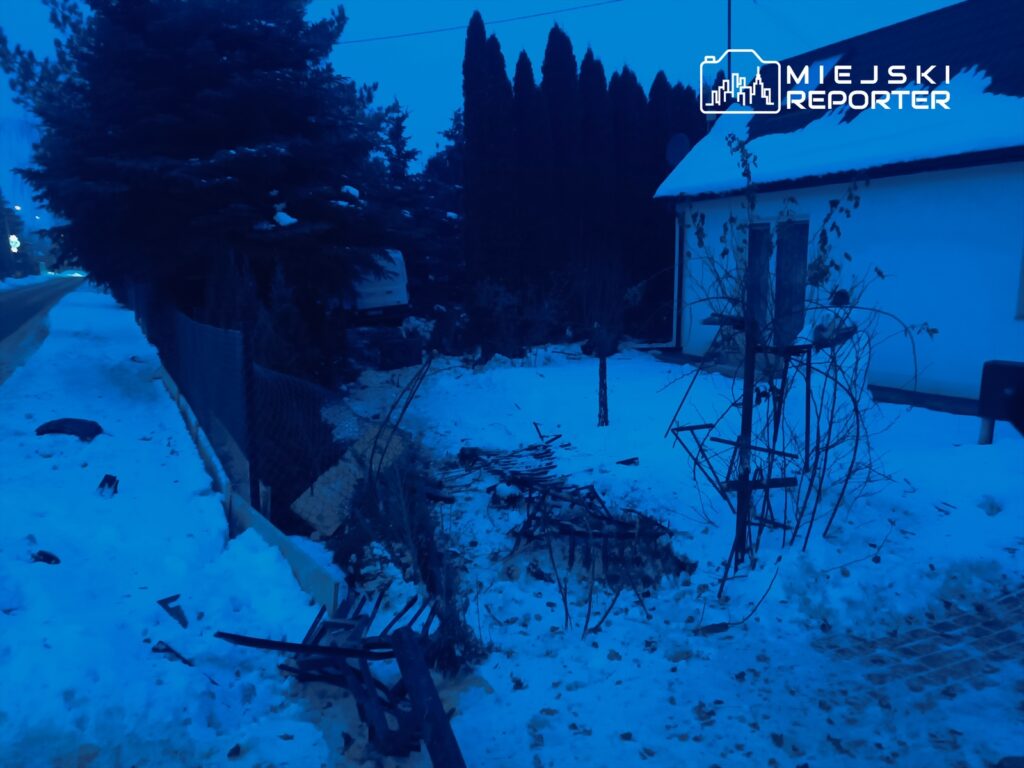 Zasypana śniegiem posesja z zniszczonym ogrodzeniem, obok leżące resztki drewna i gałęzi.