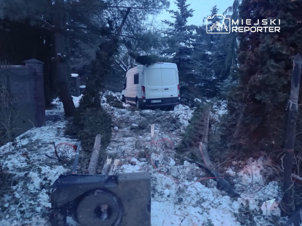 Biały furgon zaparkowany w zarośniętym ogrodzie, otoczony przez połamane gałęzie i śnieg.