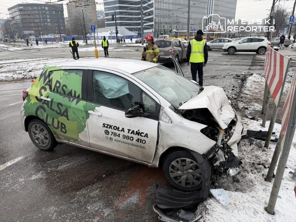 Rozbity samochód z logo "Warsaw Salsa Club" stoi na skrzyżowaniu, obok strażaków i policjantów zabezpieczających miejsce zdarzenia.