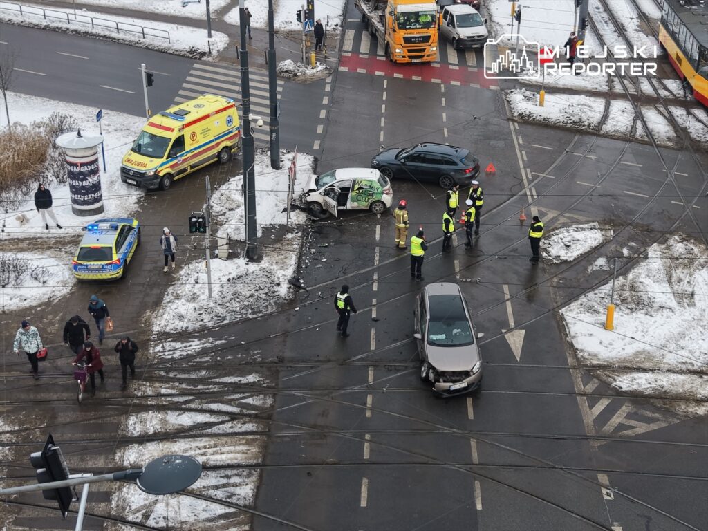 Grupa policjantów oraz strażaków bada miejsce wypadku drogowego na skrzyżowaniu, obok uszkodzonego samochodu.