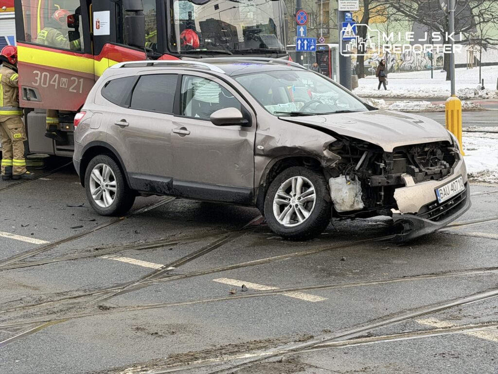 Uszkodzony samochód osobowy stojący na skrzyżowaniu z widocznymi śladami po wypadku, obok strażacy w akcji.