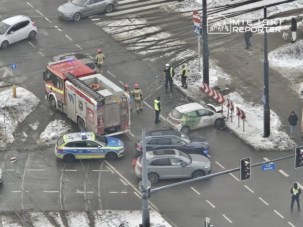 Pojazdy ratunkowe oraz policja zabezpieczają miejsce kolizji drogowej na skrzyżowaniu, w otoczeniu śniegu i ruchu ulicznego.