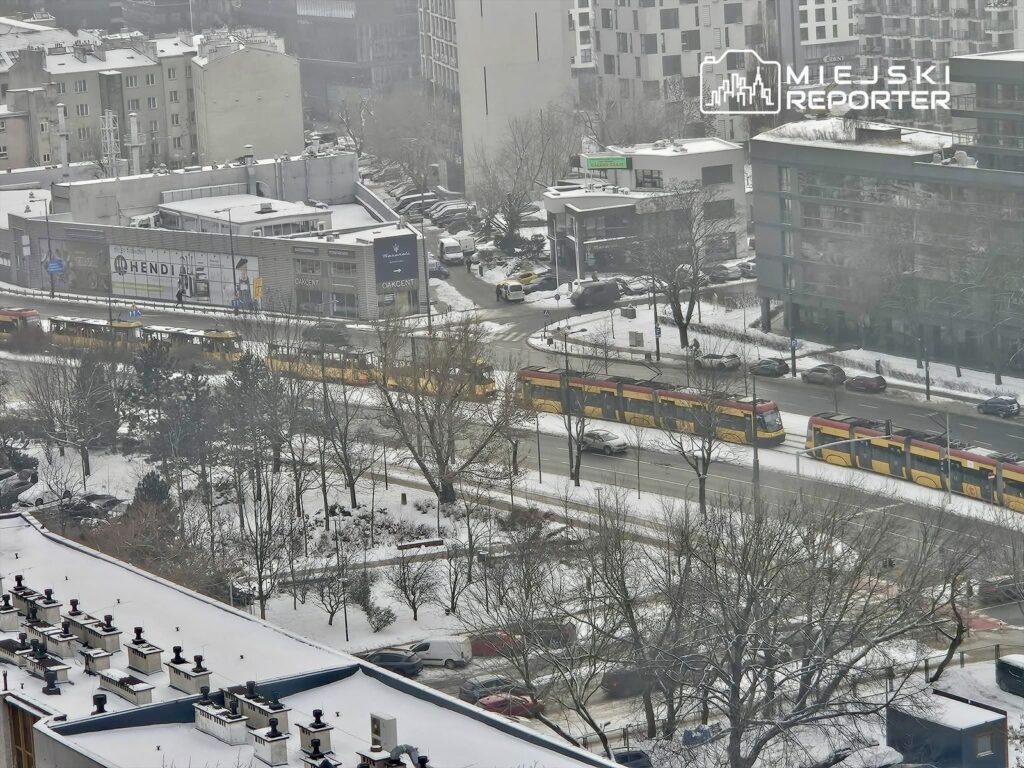 Tramwaje wjeżdżające na przystanek w zimowej scenerii z pokrytymi śniegiem ulicami i budynkami w tle.