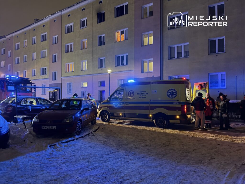 Zespół ratowników medycznych rozmawia z osobami stojącymi obok karetki w oświetlonym osiedlu mieszkaniowym.