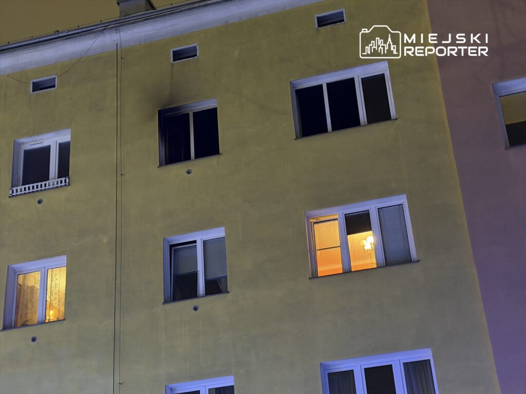 Cztery okna w żółtej elewacji budynku, jedno z włączoną lampą, w nocy.