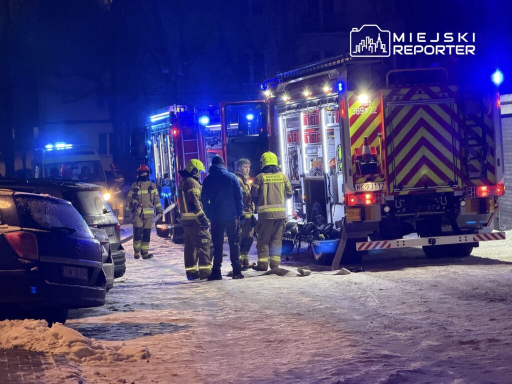 Grupa strażaków w mundurach omawia sytuację obok wozu strażackiego w oświetlonej nocą okolicy.