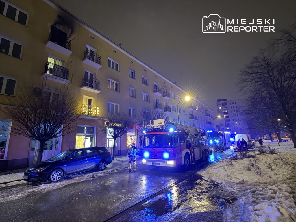 Strażacy pracują przy wozie strażackim na oświetlonej ulicy, obok budynku z zadymionym balkonem.