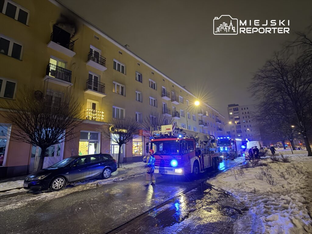 Strażacy z wozem gaśniczym interweniują przy płonącym balkonie na czwartym piętrze budynku mieszkalnego.