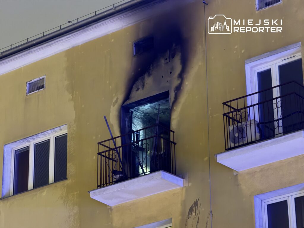 Czarna plama na elewacji budynku wskazuje na skutki pożaru, widoczny zniszczony balkon i otwarte okno.