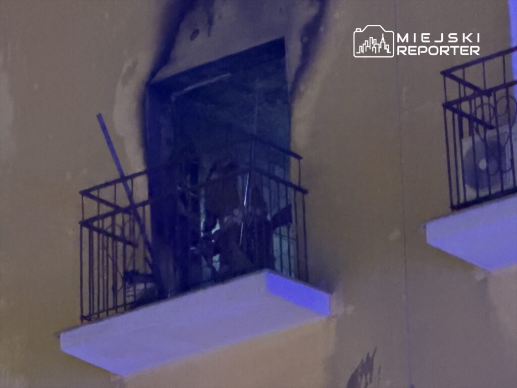 Zniszczony balkon z metalową balustradą, obok widoczne ślady po pożarze oraz uszkodzone okno.