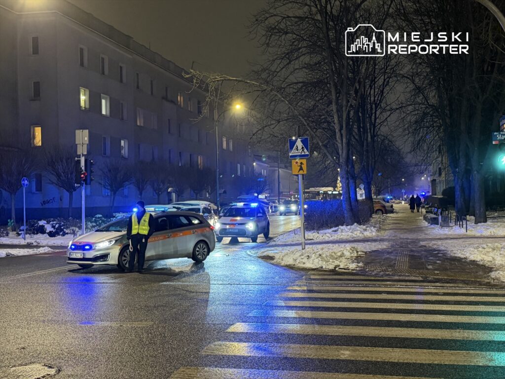 Policjant w żółtej kamizelce kieruje ruchem na oświetlonej ulicy, obok zaparkowane radiowozy z niebieskimi światłami.
