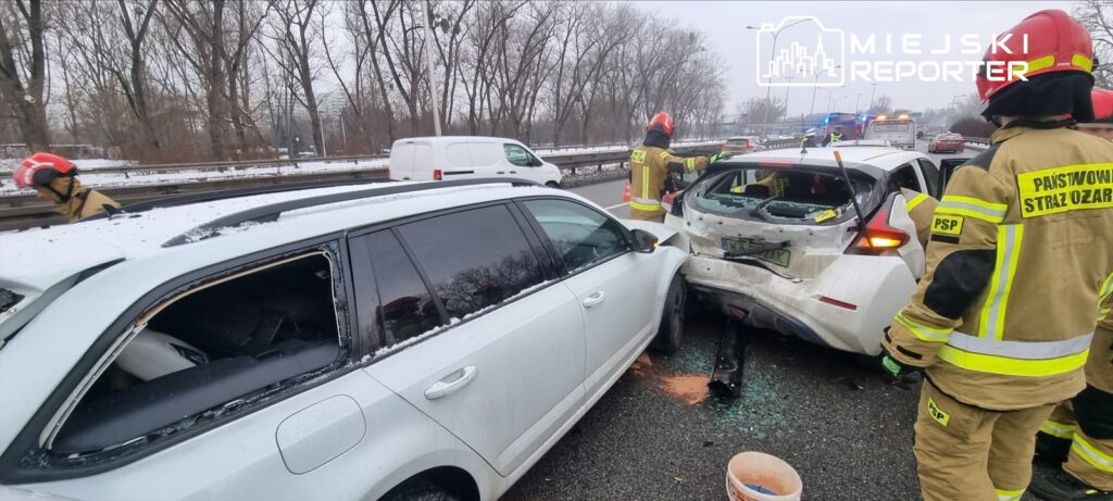 Dwa uszkodzone pojazdy po kolizji na drodze, obok pracują strażacy w mundurach i kaskach.
