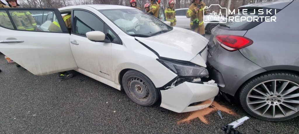 Biały samochód osobowy z uszkodzoną przednią częścią stoi na drodze, obok grupy strażaków w mundurach.