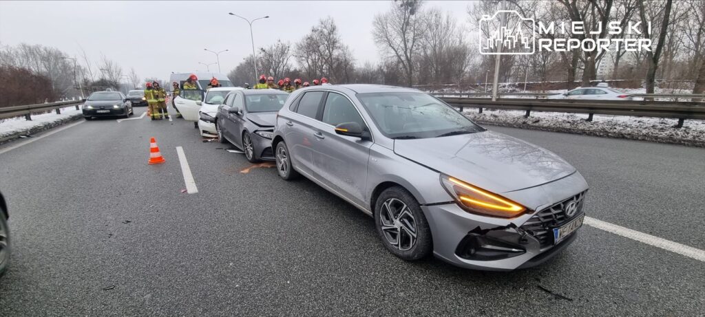 Trzy uszkodzone samochody na drodze, wokół pracujący strażacy oraz ustawione trójkąty ostrzegawcze.