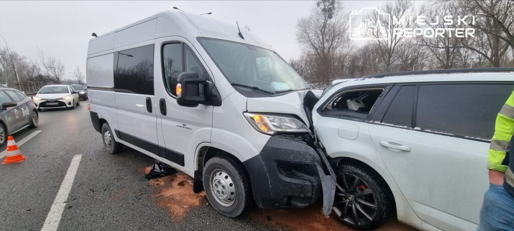 Dwa pojazdy, biały furgon i osobowy samochód, zderzone na drodze, wokół stoją inne samochody i znaki ostrzegawcze.