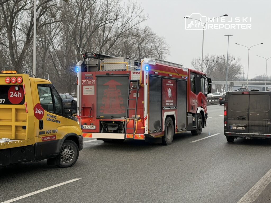 Czerwony wóz strażacki z włączonymi sygnałami świetlnymi stoi na drodze obok żółtej pomocy drogowej.