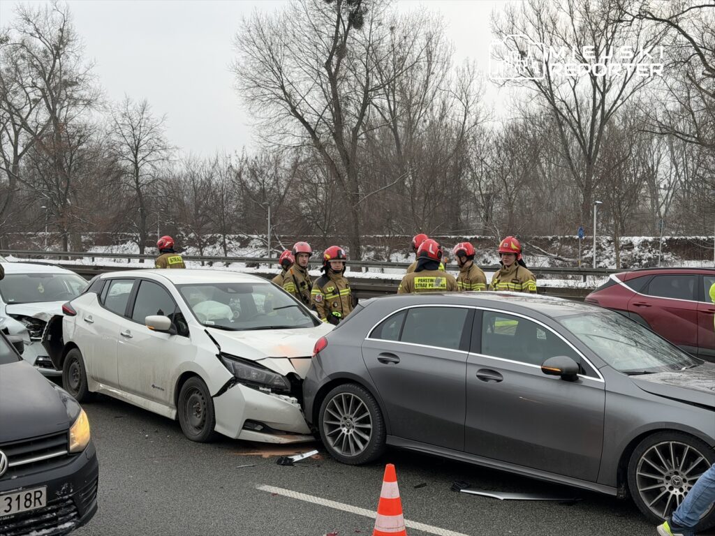 Grupa strażaków w hełmach omawia sytuację przy zderzeniu trzech samochodów na drodze, w tle widoczne drzewa.