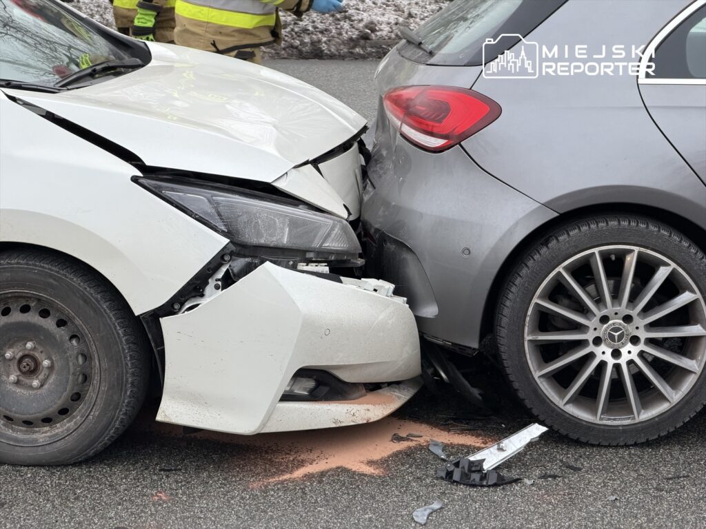 Dwa uszkodzone samochody osobowe stoją na drodze po zderzeniu, wokół nich pracują strażacy.