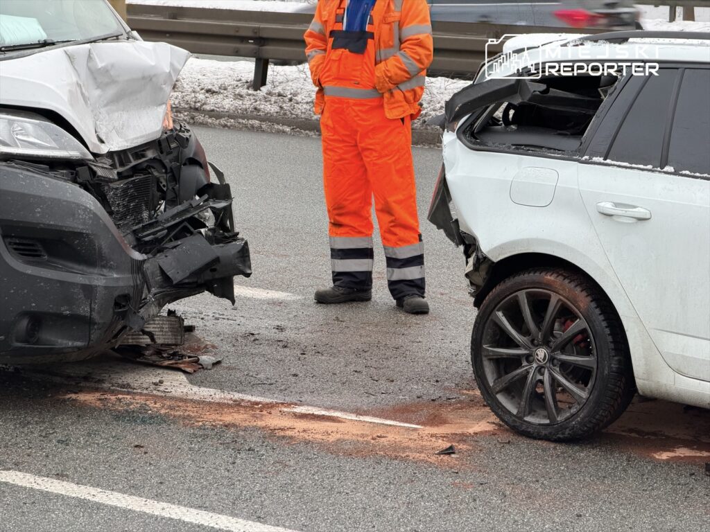Dwa uszkodzone samochody osobowe stoją na drodze, obok nich mężczyzna w pomarańczowym uniformie.