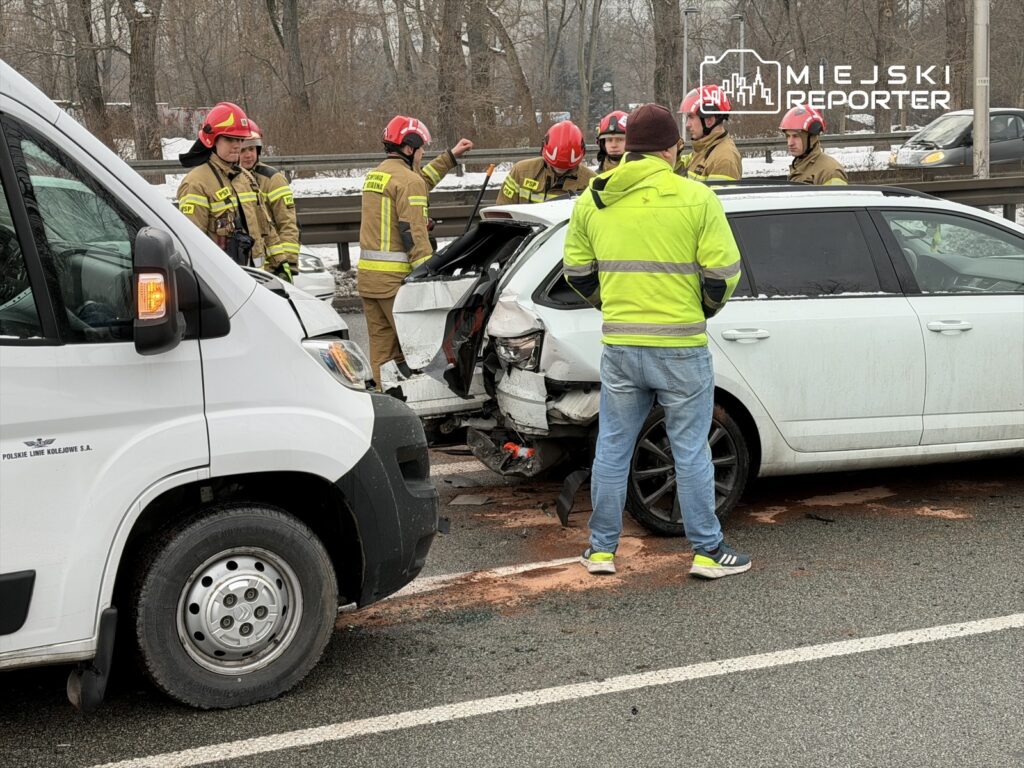 Grupa strażaków w kaskach omawia wypadek drogowy obok uszkodzonego samochodu na drodze.