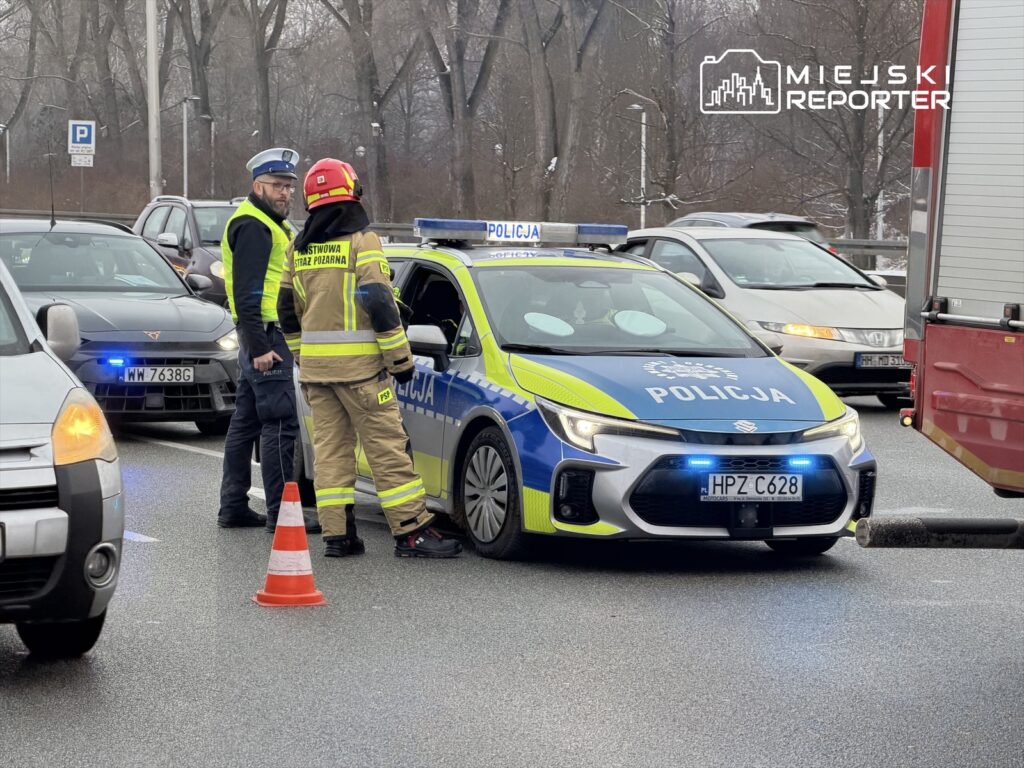 Policjant i strażak rozmawiają obok radiowozu na ruchliwej drodze, otoczeni przez samochody.