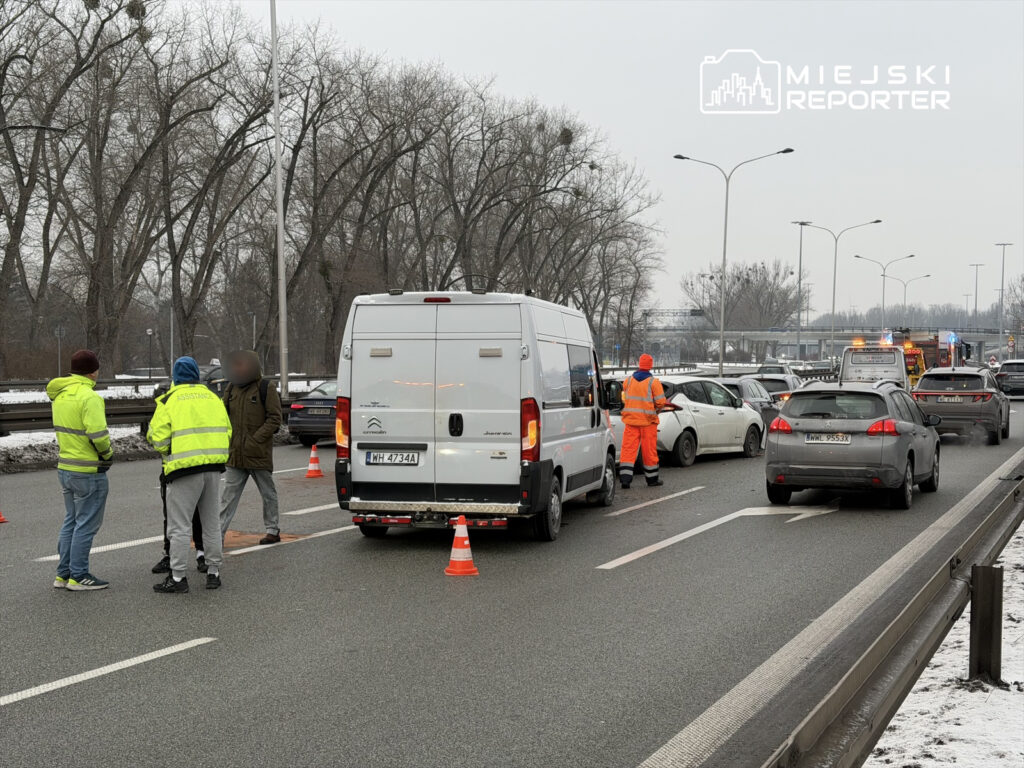 Grupa osób w odblaskowych kamizelkach rozmawia obok uszkodzonych samochodów na zatłoczonej drodze w zimowej scenerii.