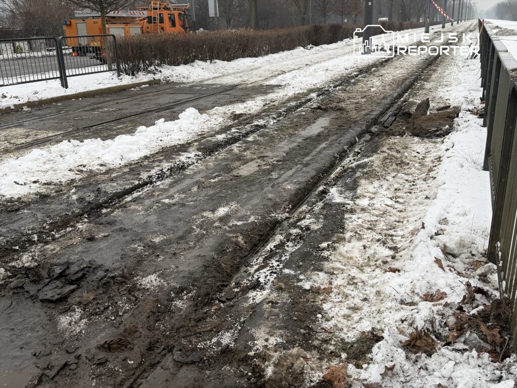 Zaniedbane torowisko tramwajowe pokryte błotem i śniegiem, w tle pomarańczowy pojazd budowlany.