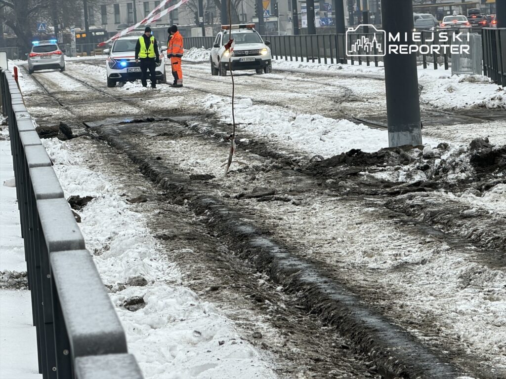 Na zaśnieżonej ulicy pracownicy w odblaskowych kamizelkach rozmawiają obok uszkodzonego torowiska tramwajowego.