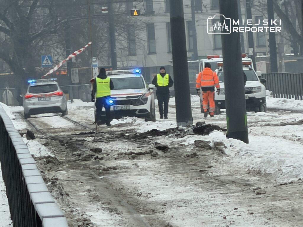 Grupa pracowników w odblaskowych kamizelkach zabezpiecza teren budowy w zaśnieżonej okolicy, obok zaparkowanych radiowozów.