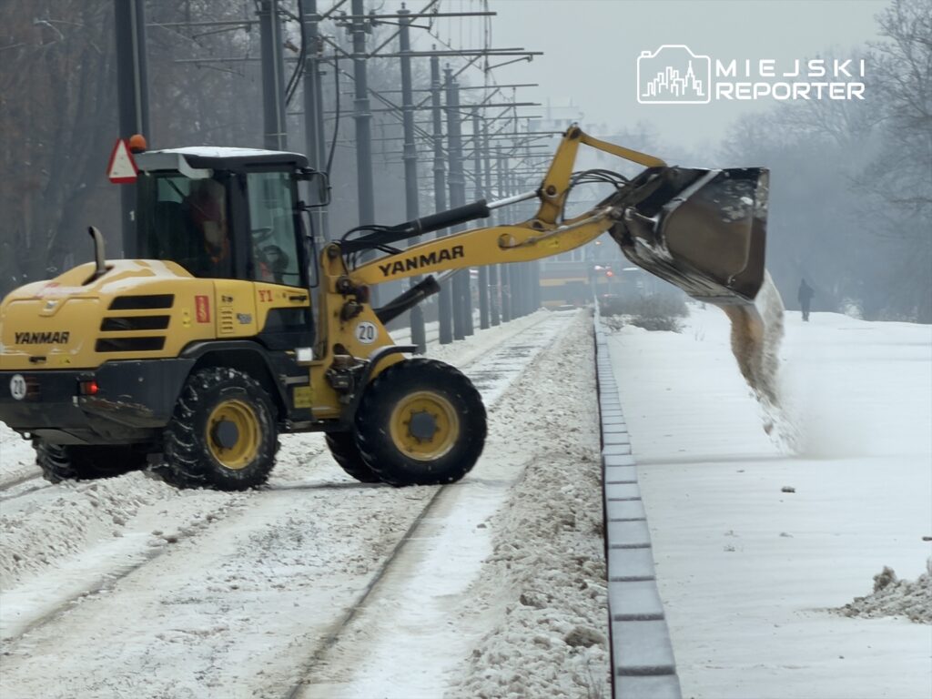 Maszyna budowlana z łyżką odśnieżającą usuwa śnieg z torów tramwajowych w zimowej scenerii.