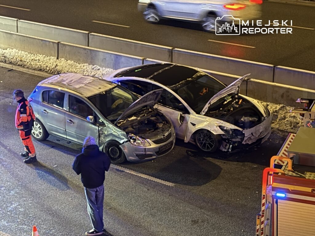 Dwa uszkodzone samochody stoją na drodze, a ratownik medyczny w czerwonym stroju obserwuje sytuację.