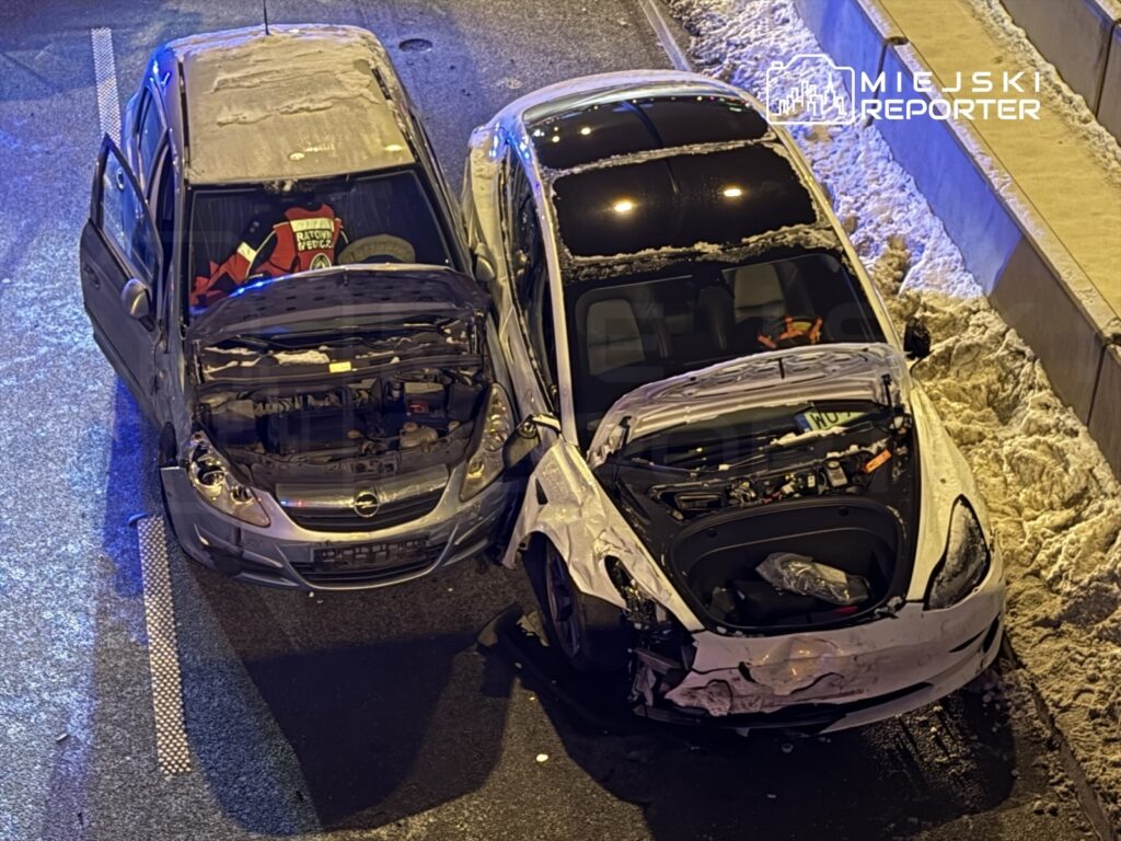 Dwa samochody osobowe z otwartymi maskami, jeden z uszkodzeniami przodu, stoją na oświetlonej drodze.