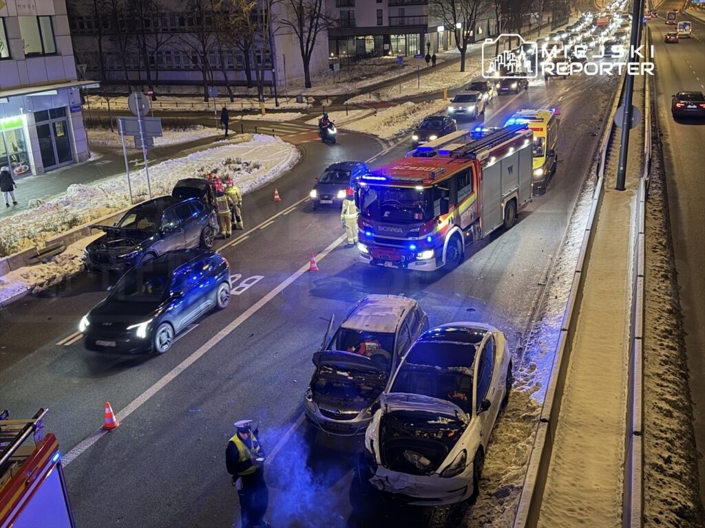 W nocy na ulicy widoczne są dwa uszkodzone samochody, obok stoi wóz strażacki, a strażacy udzielają pomocy.